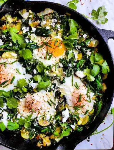 A skillet filled with vibrant Green Shakshuka, featuring wilted greens, perfectly cooked eggs with runny yolks, and garnished with fresh herbs like parsley and dill. Served with crusty bread on the side for scooping.